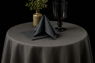 KI-generiert. Ein runder Tisch mit grauer Tischdecke, gefalteter dunkler Serviette, Kelchglas und einer kleinen Vase mit Zweigen.